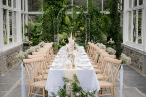 The glasshouse at Vandeleur Walled Garden & Visitor Centre with a long table set up for a special event. White table cloth, candles, table settings, and beautiful floral arrangements.