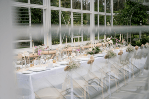 The glasshouse at Vandeleur Walled Garden & Visitor Centre with a long table set up for a special event viewed from the outside through a window. White table cloth, candles, table settings, and beautiful floral arrangements.