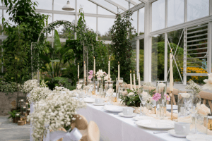 The glasshouse at Vandeleur Walled Garden & Visitor Centre with a long table set up for a special event. White table cloth, candles, table settings, and beautiful floral arrangements. Closeup shot with the glasshouse flower bed in the background.