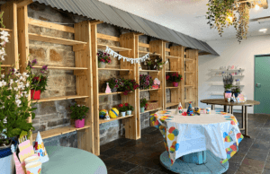 Image of the Woodland room at Vandeleur Walled Garden. The room is long, with stone floors and wooden shelving on the left-hand side. There are three tables dressed up for a party with birthday decorations and flowers. A couple of "Happy Birthday" signs hang from the shelves, which are also filled with flowers and a few toys.